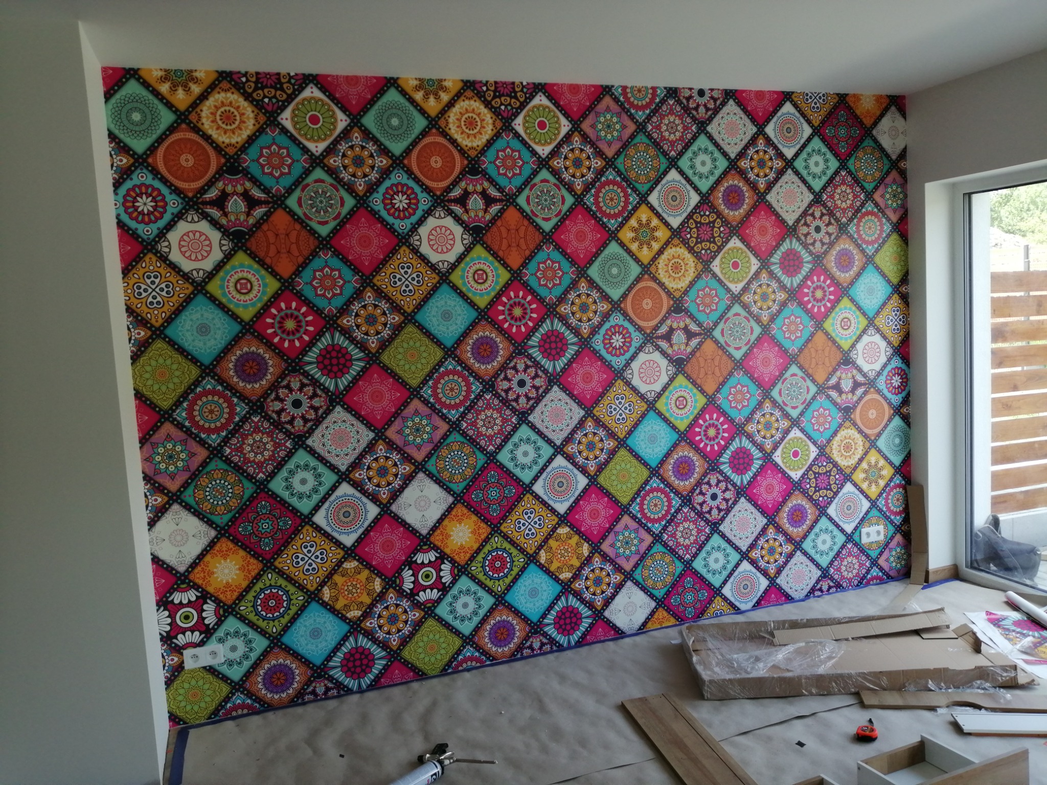 Ściana pokryta kolorową tapetą w geometryczne wzory, częściowo zabezpieczona kartonem i folią, widoczne narzędzia i materiały budowlane na podłodze.