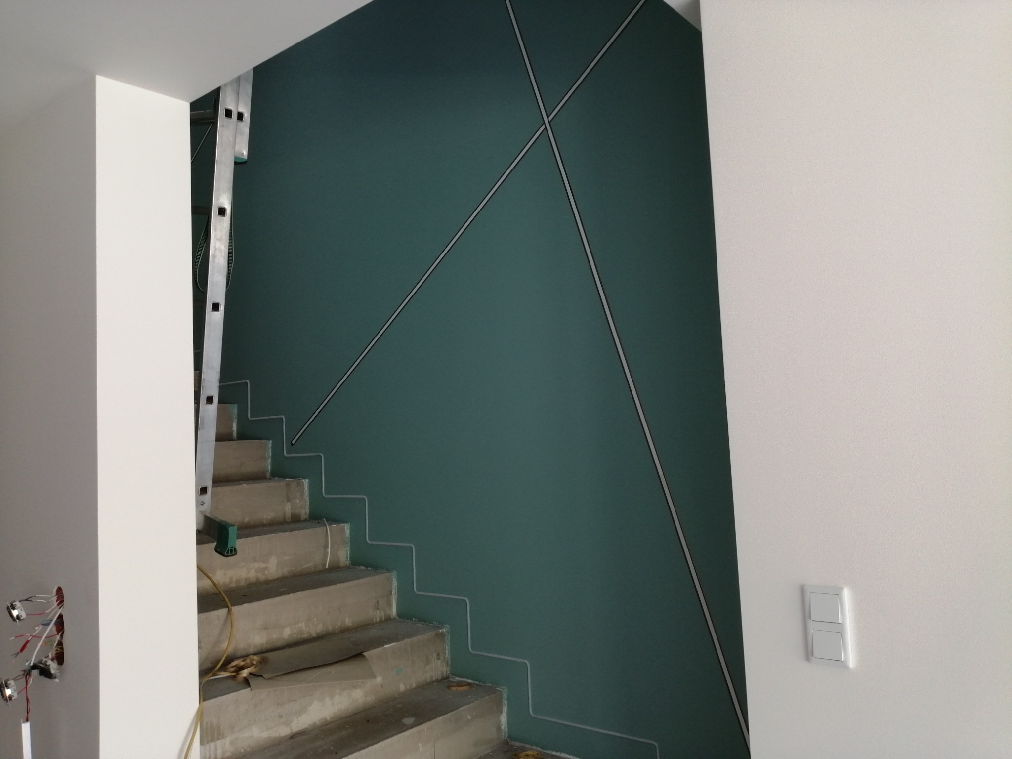 Klatka schodowa w trakcie wykańczania, ściana pomalowana na ciemnozielono z geometrycznym wzorem z listew, surowe betonowe schody zabezpieczone folią, widoczna drabina i instalacja elektryczna.