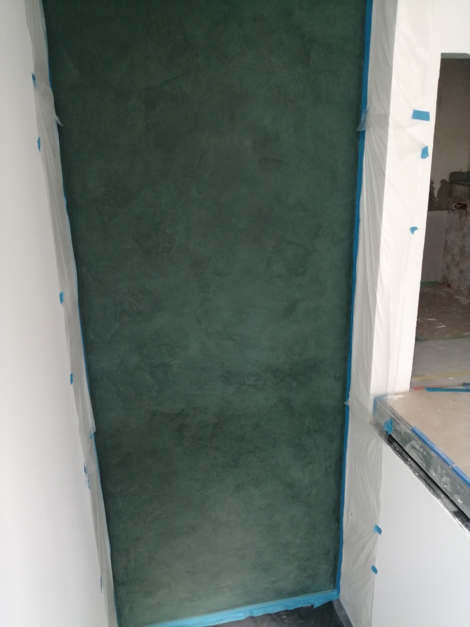 Ściana pokryta dekoracyjną, ciemnozieloną strukturą, zabezpieczona folią malarską z niebieską taśmą, w trakcie prac wykończeniowych.