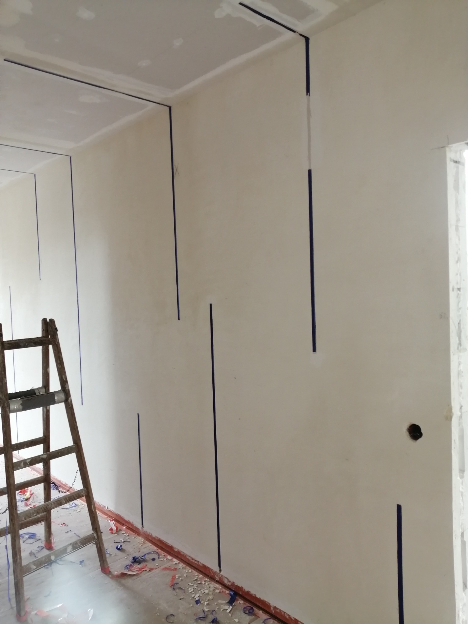 Ściana przygotowana do położenia gładzi, z pionowymi paskami taśmy malarskiej wyznaczającymi miejsca na listwy, drewniana drabina oparta o ścianę, podłoga zabezpieczona folią malarską, widoczne...