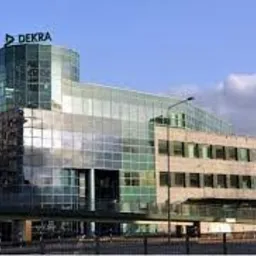 Nowoczesny budynek biurowy z przeszkloną fasadą i logo DEKRA na dachu, widoczny na tle błękitnego nieba z chmurami.