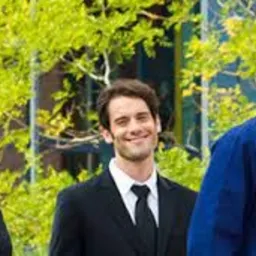 Uśmiechnięci pracownicy: kobieta w czarnym garniturze, mężczyzna w garniturze z krawatem oraz mężczyzna w niebieskiej koszuli na tle zieleni.