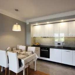 Nowoczesna, jasna kuchnia z białymi, lakierowanymi szafkami, czarnym blatem i marmurowym backsplashiem, uzupełniona stołem z nakryciem i białymi krzesłami, oświetlona lampą wiszącą z abażurem.