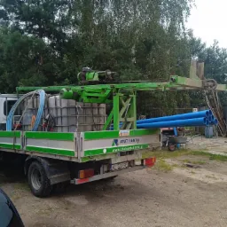 Ciężarówka z platformą wiertniczą i zbiornikiem na wodę, zaparkowana na żwirowym podłożu, z niebieskimi rurami w skrzyni ładunkowej i zielonym osprzętem wiertniczym, otoczona zielenią drzew.