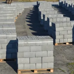 Szereg ułożonych na paletach szarych bloczków betonowych z otworami, widoczne cienie, w tle kolejne rzędy bloczków i palety.