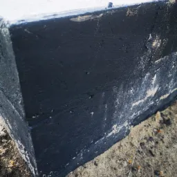 Fragment fundamentu pomalowanego czarną, hydroizolacyjną substancją, widoczne ślady po malowaniu na betonie i piasku.