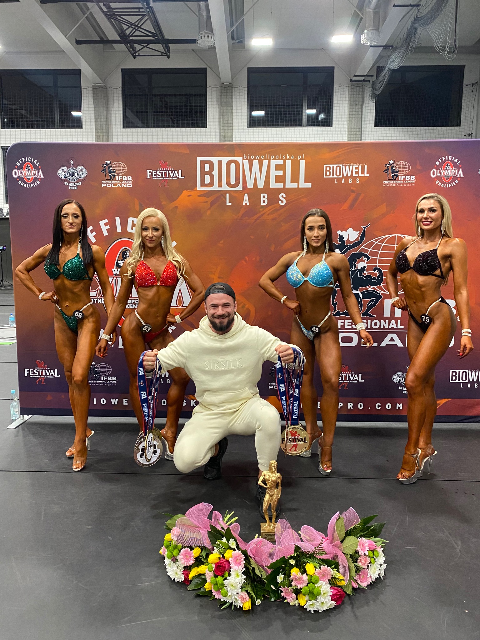 Zdjęcie z zawodów fitness: Pięć osób, w tym cztery kobiety w błyszczących bikini i mężczyzna w dresie, pozują na scenie z nagrodami i kwiatami, na tle baneru reklamowego.