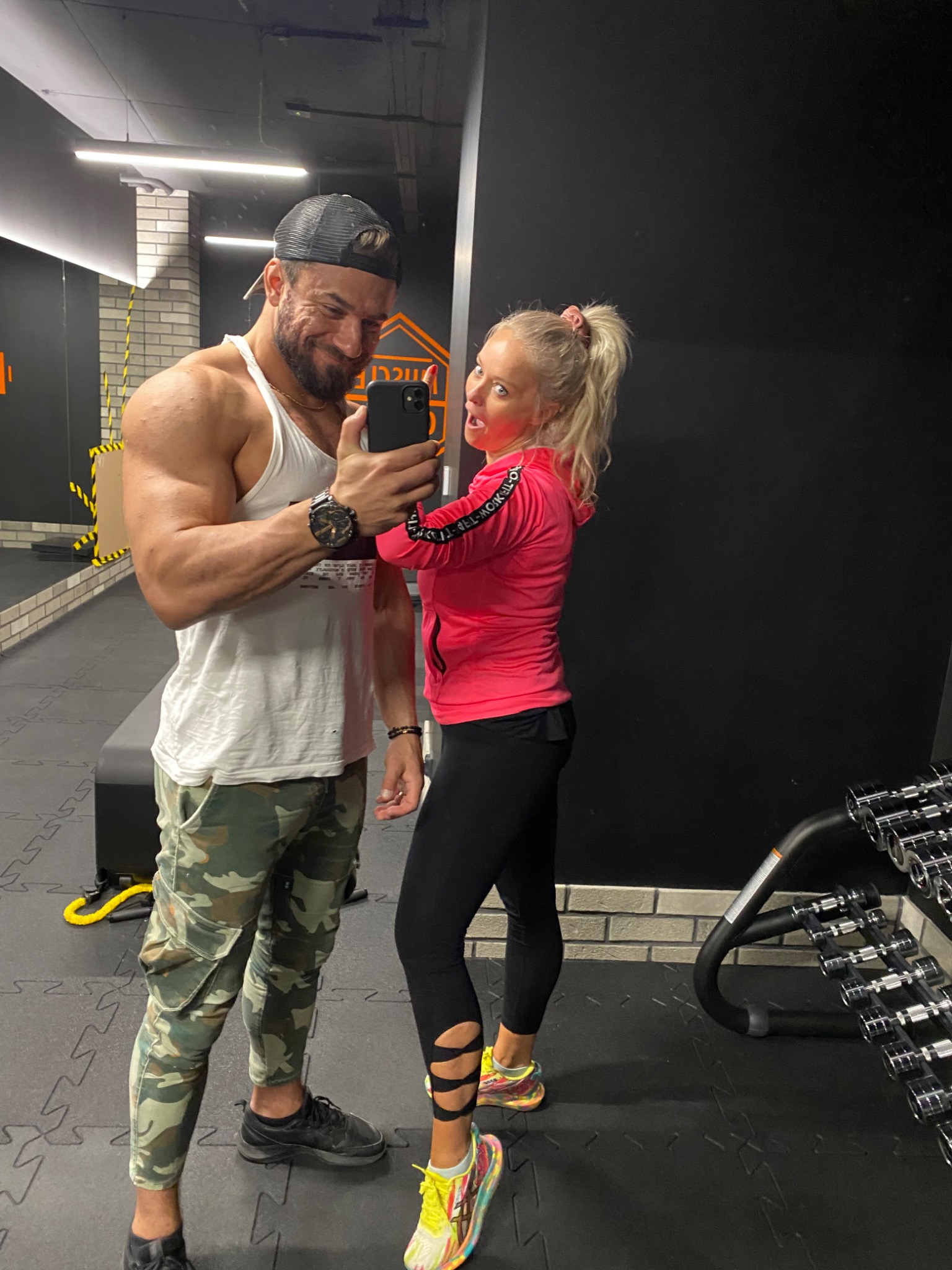 Para w siłowni robiąca selfie: mężczyzna z wyraźnie zarysowanymi mięśniami w białym podkoszulku i spodniach moro, kobieta w różowej bluzie i czarnych legginsach z wycięciami, oboje uśmiechnięci,...