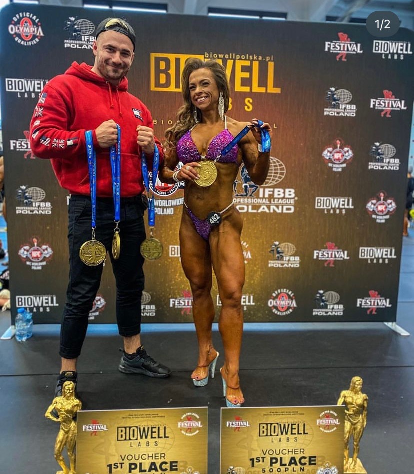 Kobieta w fioletowym bikini pozuje z mężczyzną na tle sceny po zawodach kulturystycznych, oboje trzymają złote medale i vouchery za pierwsze miejsce.