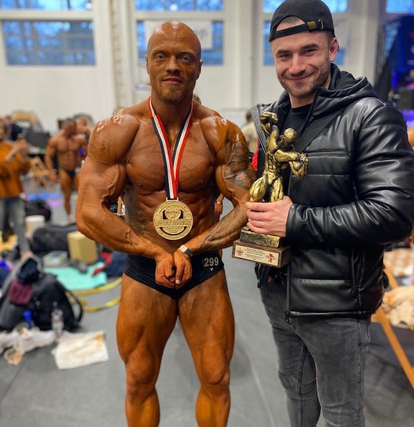 Uśmiechnięty kulturysta z medalem i numerem startowym 299, pozujący z nagrodą i mężczyzną w skórzanej kurtce. Tło sceny z innymi uczestnikami.