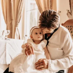 Dziecko w białej sukience i opasce na głowie, trzymane na kolanach przez osobę w białej koszuli na tle eleganckiego wnętrza z zasłonami i nakrytym stołem.