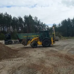 Żółta koparko-ładowarka New Holland na piaszczystym placu budowy, w tle ciężarówka z przyczepą i las iglasty pod pochmurnym niebem.