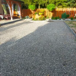 Szara nawierzchnia z drobnego kruszywa na posesji, widoczny cień drzewa, drewniany płot i altana w tle.