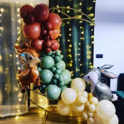 Dekoracja balonowa na metalowym stelażu w kształcie koła, z balonami w odcieniach brązu, zieleni i beżu, uzupełniona balonami foliowymi w kształcie sarenki i zajączka, na tle zasłon z lampkami.