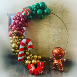 Dekoracja balonowa w kształcie okręgu z motywami świątecznymi: zielone, czerwone i złote balony, lizak, piernikowy ludzik i prezenty w czerwonych pudełkach.