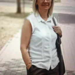 Kobieta w okularach, białej koszuli i krótkich spodenkach, stojąca na chodniku z torbą na ramieniu.