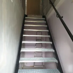 Wnętrze w trakcie remontu z widokiem na metalowe schody kratowe z czarną balustradą, w tle rozstawiona drabina.