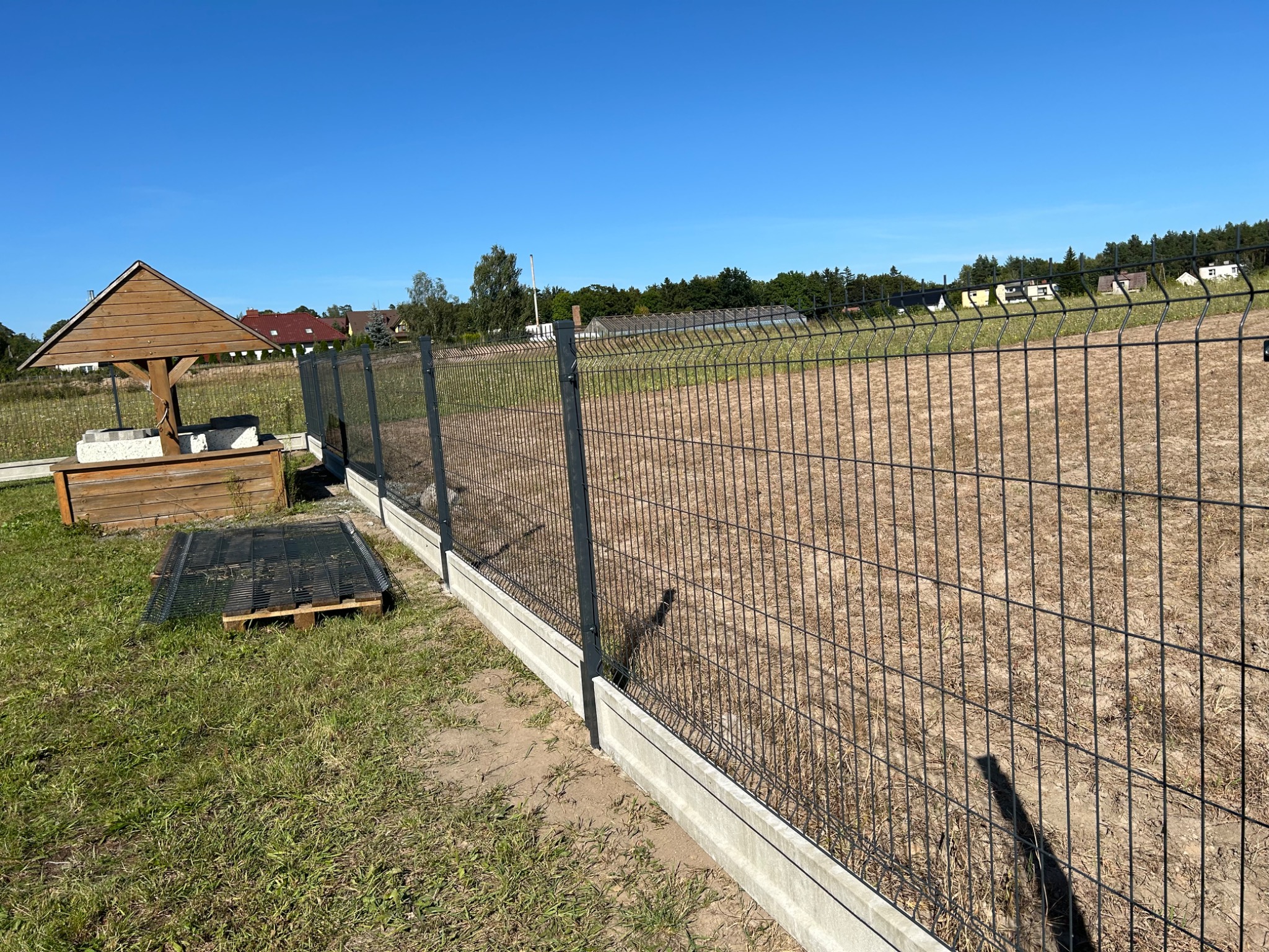 Nowoczesne ogrodzenie panelowe z podmurówką betonową na działce, widoczna studnia z drewnianą konstrukcją w tle, słoneczny dzień.