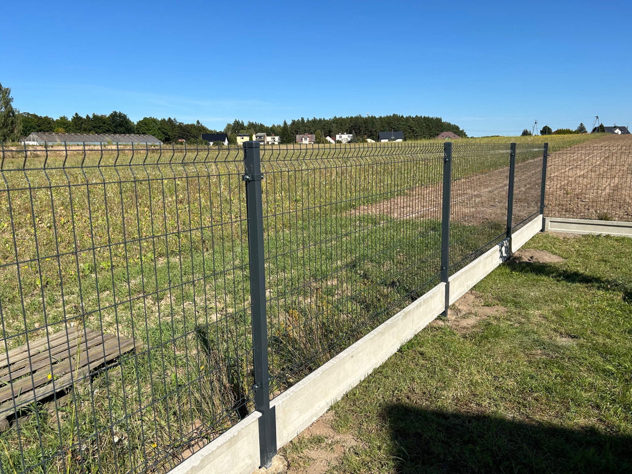 Nowoczesne ogrodzenie panelowe z podmurówką betonową na tle zielonego pola i zabudowań w oddali, wykonane w słoneczny dzień.