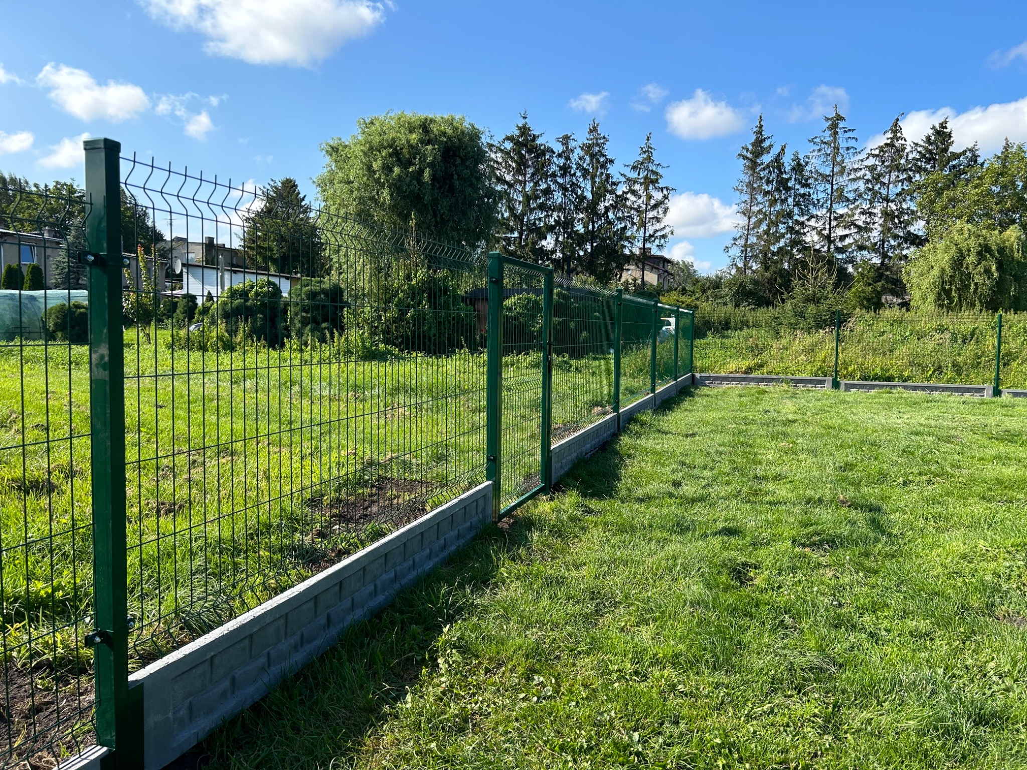 Zielone ogrodzenie panelowe z podmurówką z szarego betonu na tle zielonego trawnika i drzew pod błękitnym niebem.