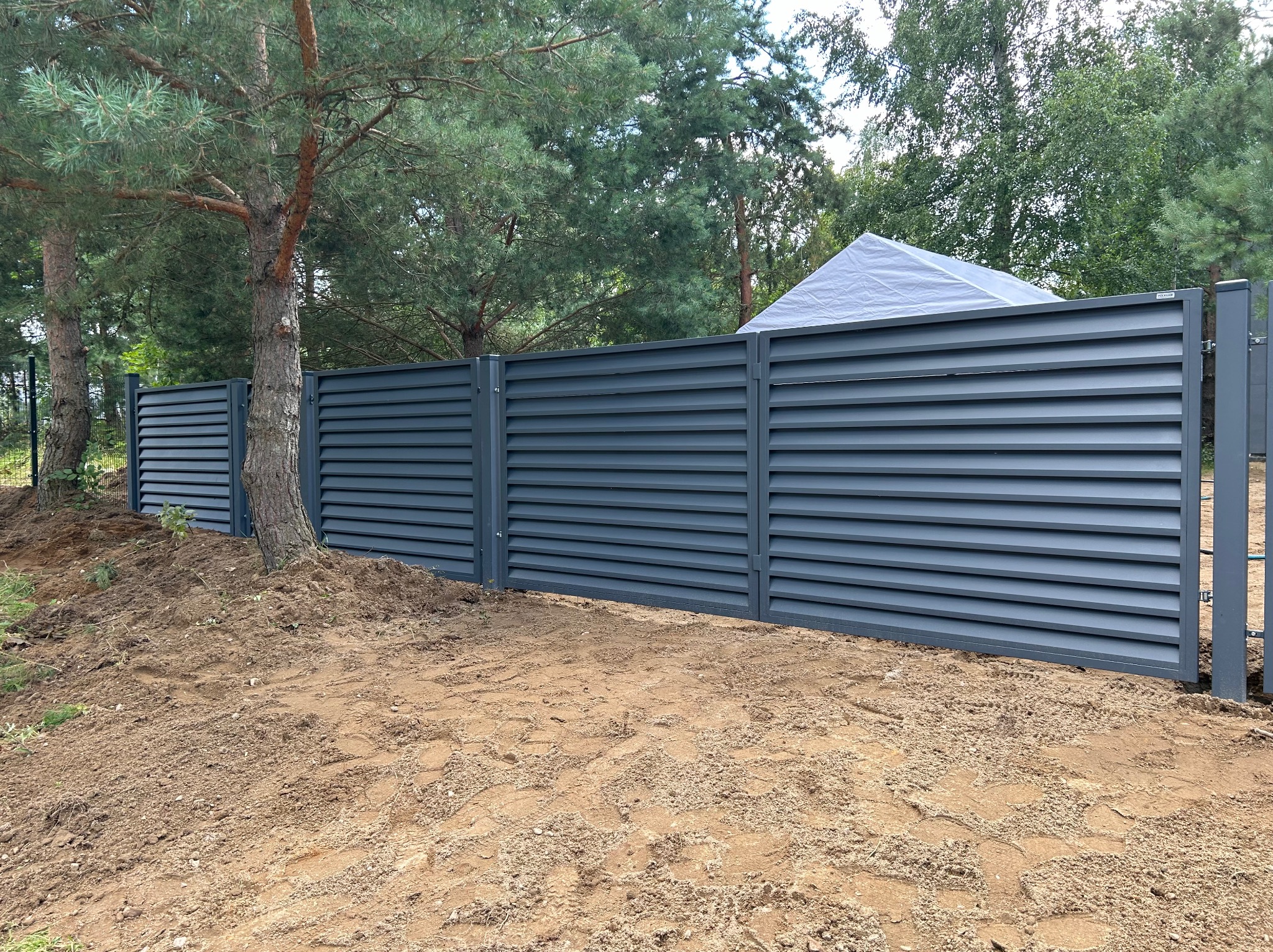 Nowoczesne, poziome ogrodzenie panelowe w kolorze grafitowym, zamontowane w Kartuzach, z widocznymi śladami prac ziemnych i drzewem wkomponowanym w linię ogrodzenia.