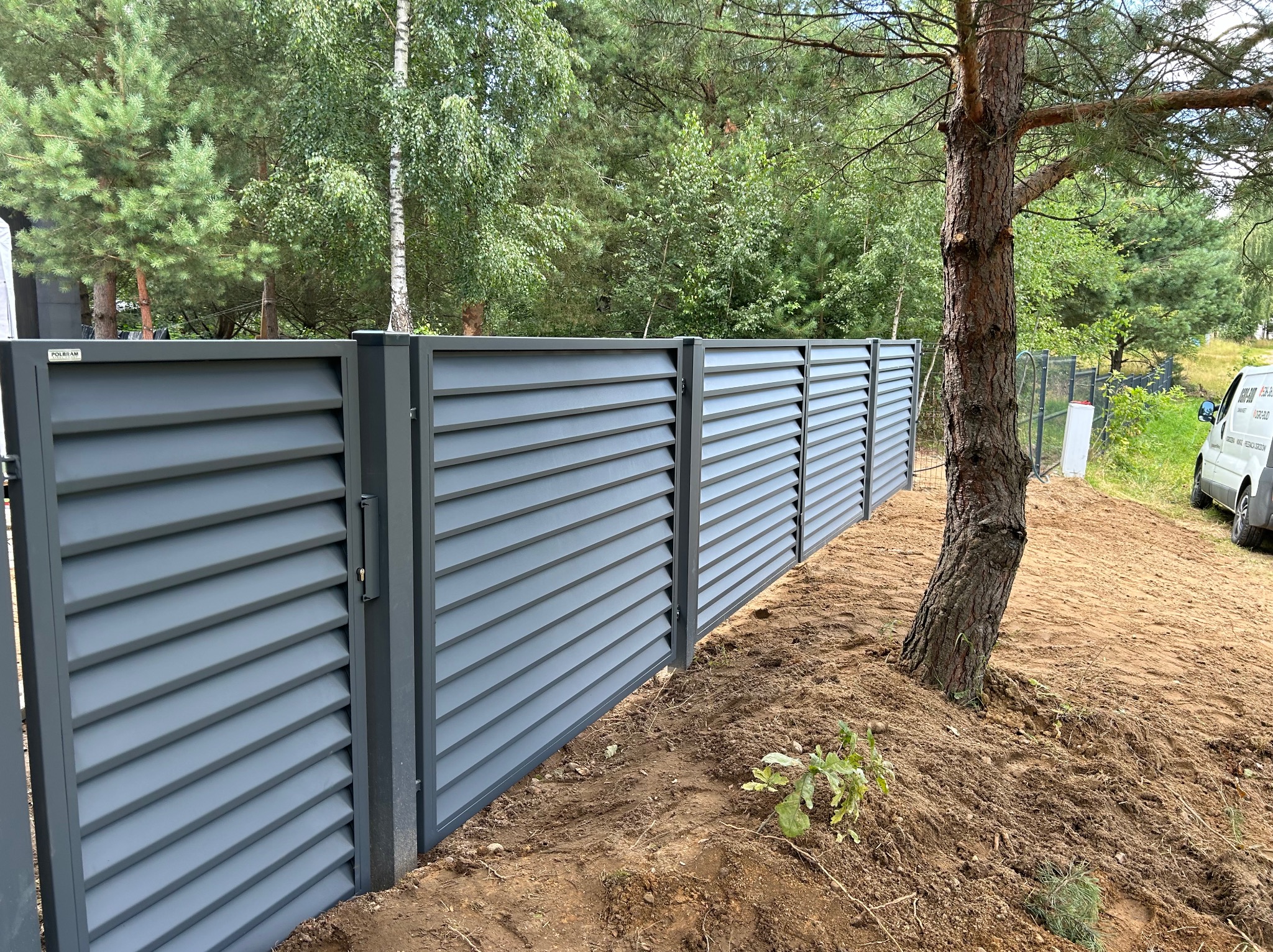 Nowoczesne, szare ogrodzenie panelowe z poziomymi lamelami, widoczne na tle zieleni drzew, z ziemią po montażu i fragmentem białego samochodu dostawczego w tle.