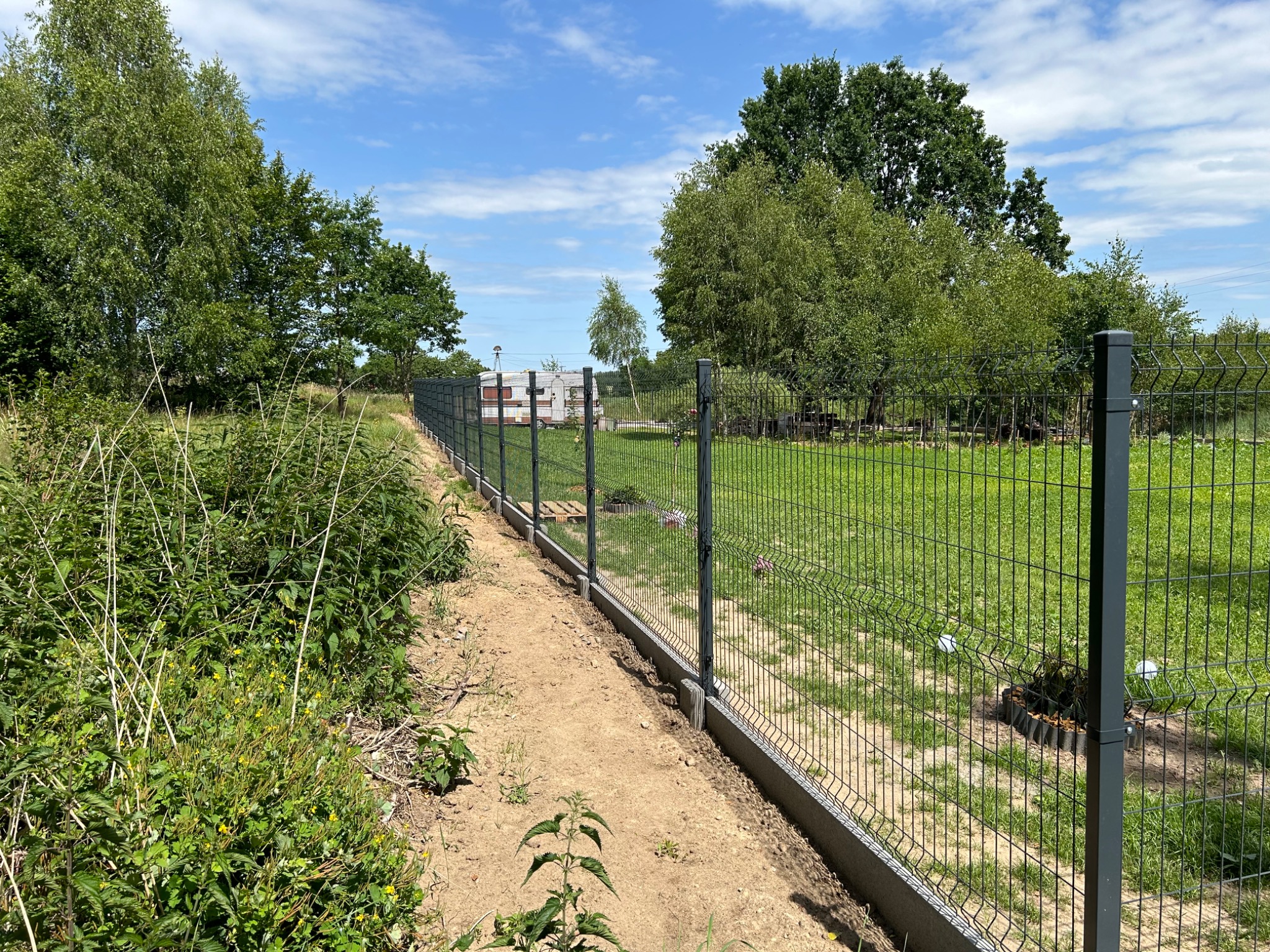 Nowo zamontowane, szare ogrodzenie panelowe z podmurówką betonową, widok z boku na tle zielonego trawnika i drzew w słoneczny dzień.