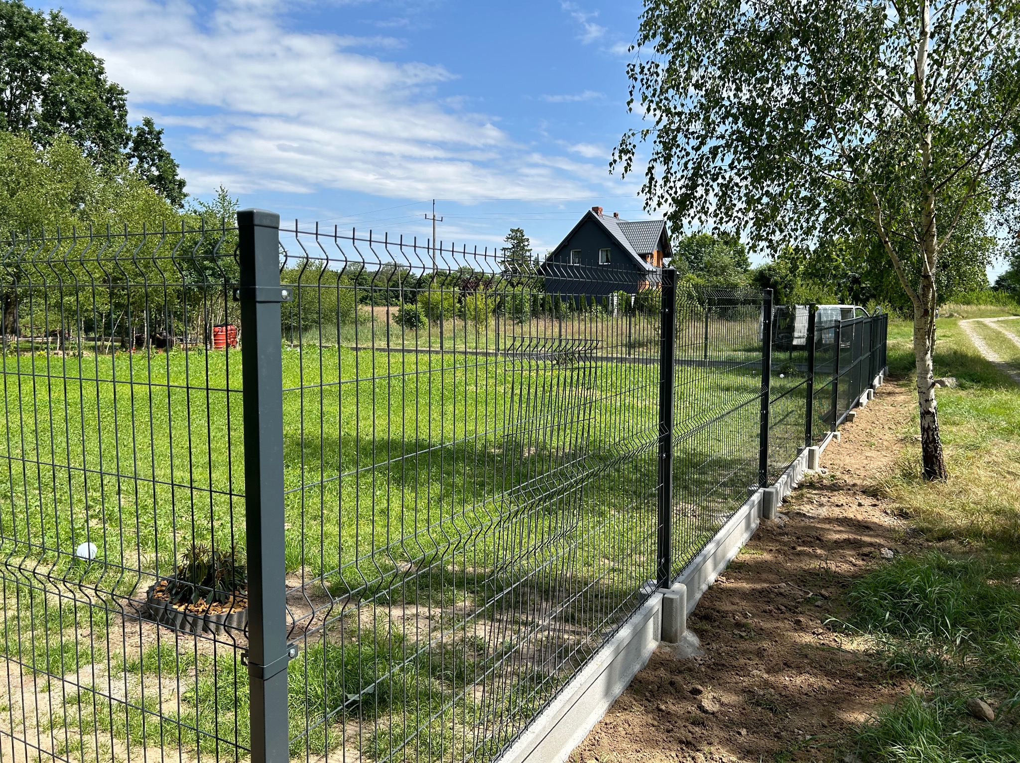 Nowo postawione, ciemne ogrodzenie panelowe z podmurówką betonową otaczające zielony trawnik, widok na dom w oddali i boczna, polna droga.