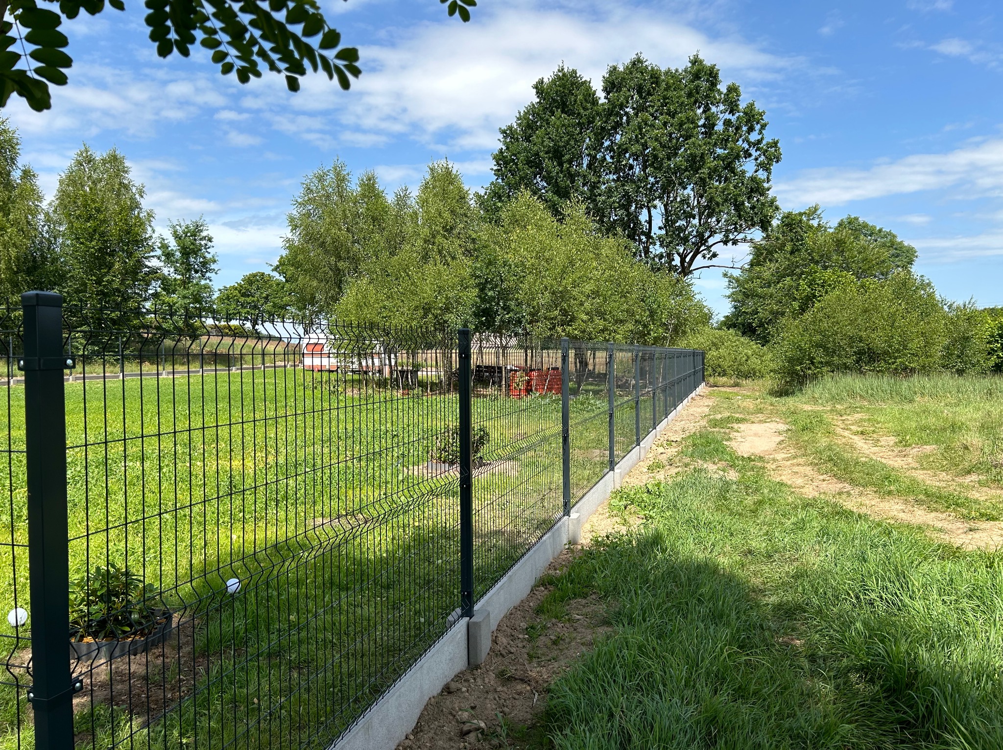 Nowoczesne ogrodzenie panelowe z podmurówką betonową, widok z boku, zielony trawnik i drzewa w tle, słoneczny dzień.