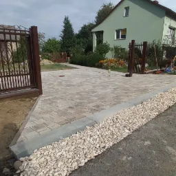 Ułożona kostka brukowa na podjeździe z obrzeżami wypełnionymi białym grysem, widok na dom z zieloną elewacją i metalową bramą w tle.