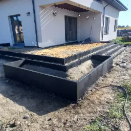 Wykonane z czarnych element&oacute;w obrzeża rabat w ogrodzie przy nowoczesnym domu, wypełnione piaskiem, widoczne ślady prac ziemnych.