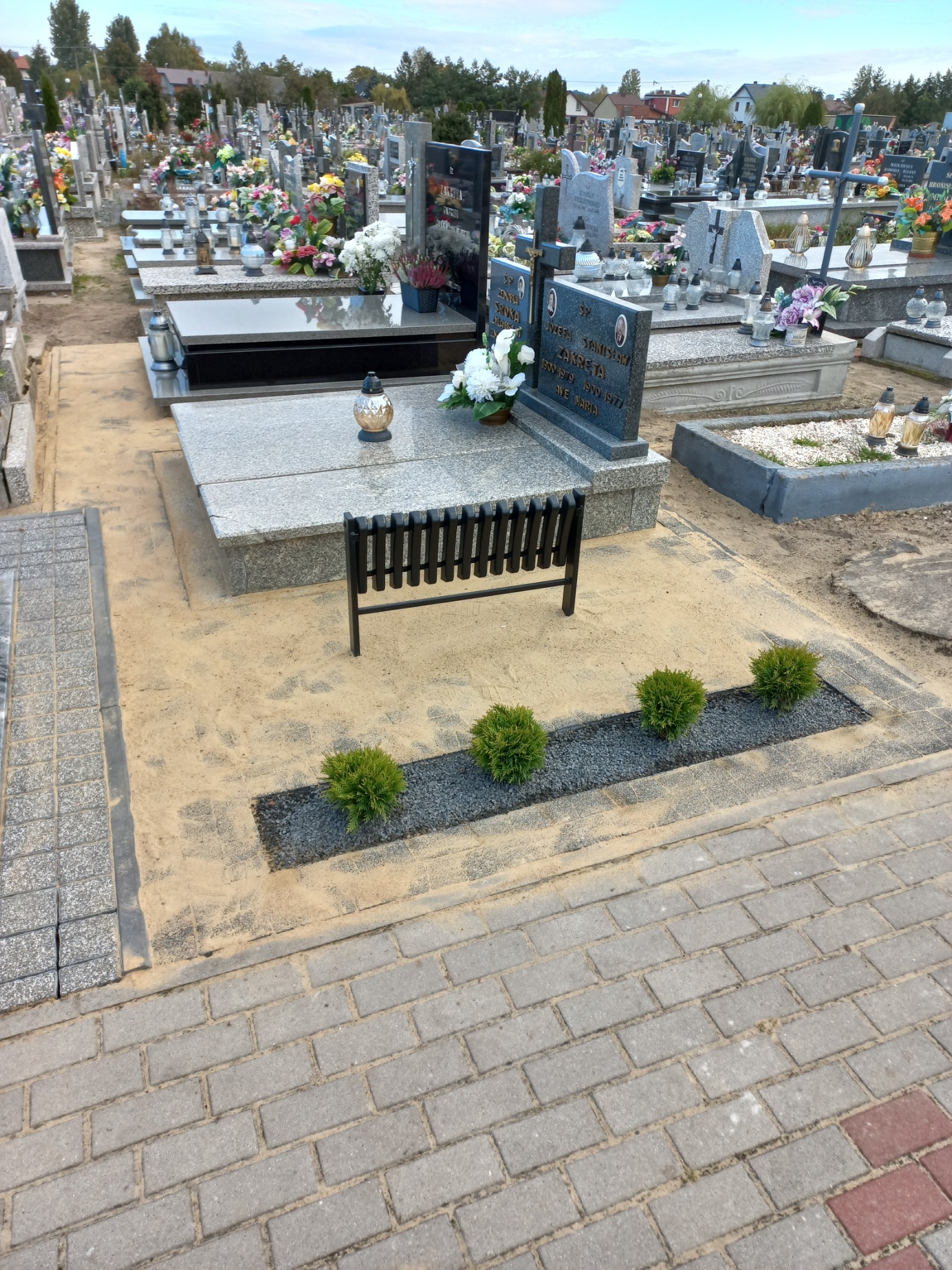 Wykończone miejsce pochówku z szarą kostką brukową, żwirem, czterema małymi krzewami i czarną ławką na cmentarzu, widok z góry.