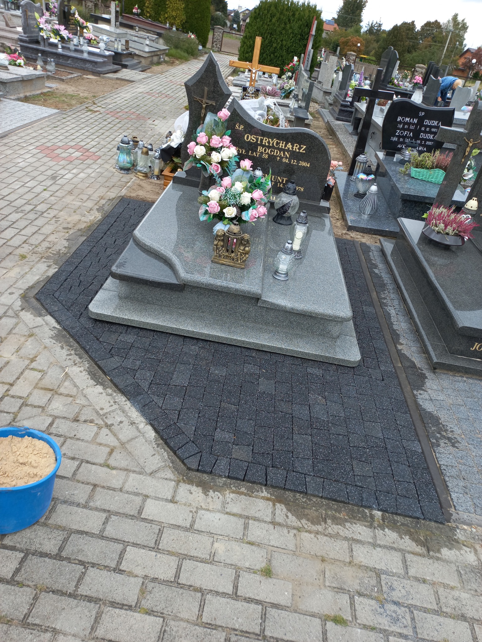 Ciemna kostka brukowa ułożona wokół nagrobka z kwiatami i zniczami, widoczny niebieski pojemnik z piaskiem, cmentarz w tle.