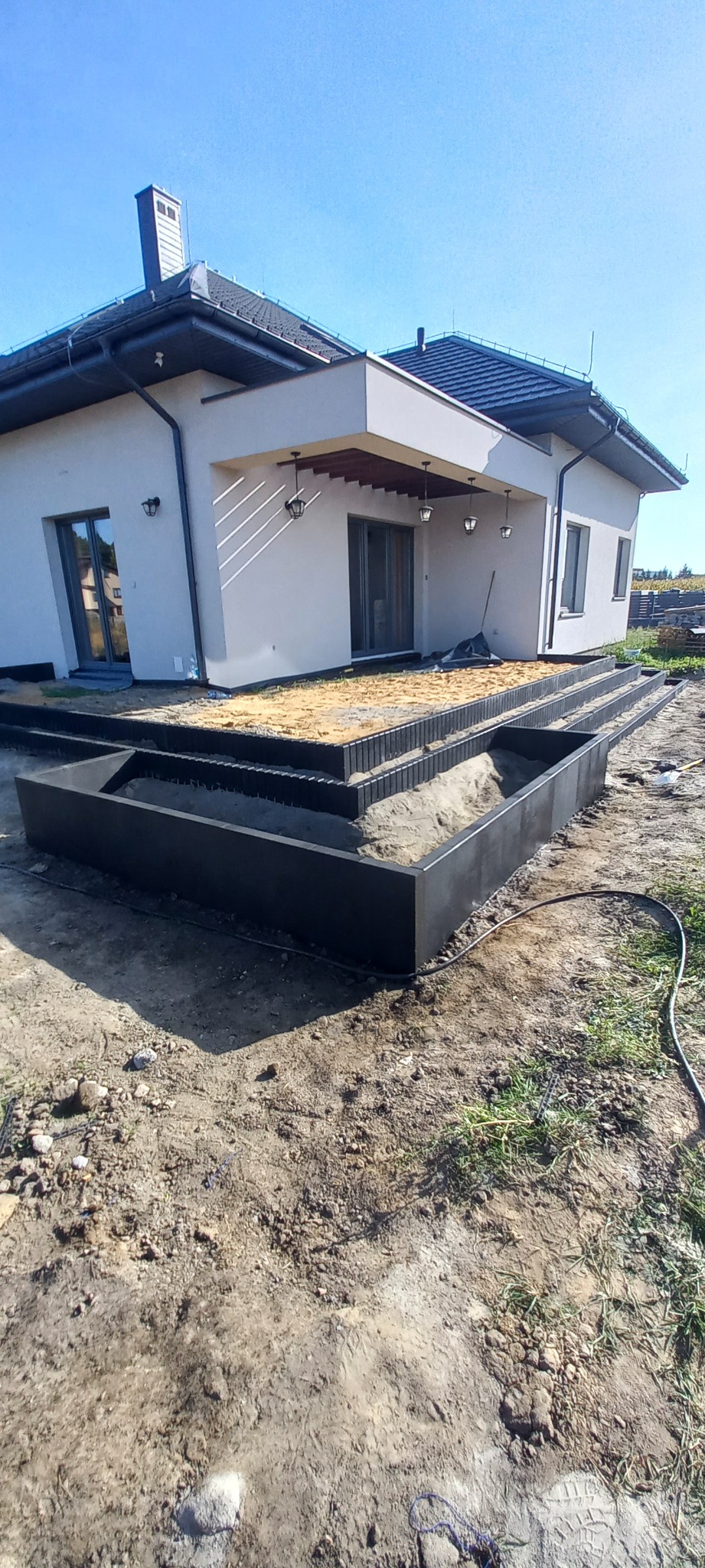 Wykonane z czarnych elementów obrzeża rabat w ogrodzie przy nowoczesnym domu, wypełnione piaskiem, widoczne ślady prac ziemnych.