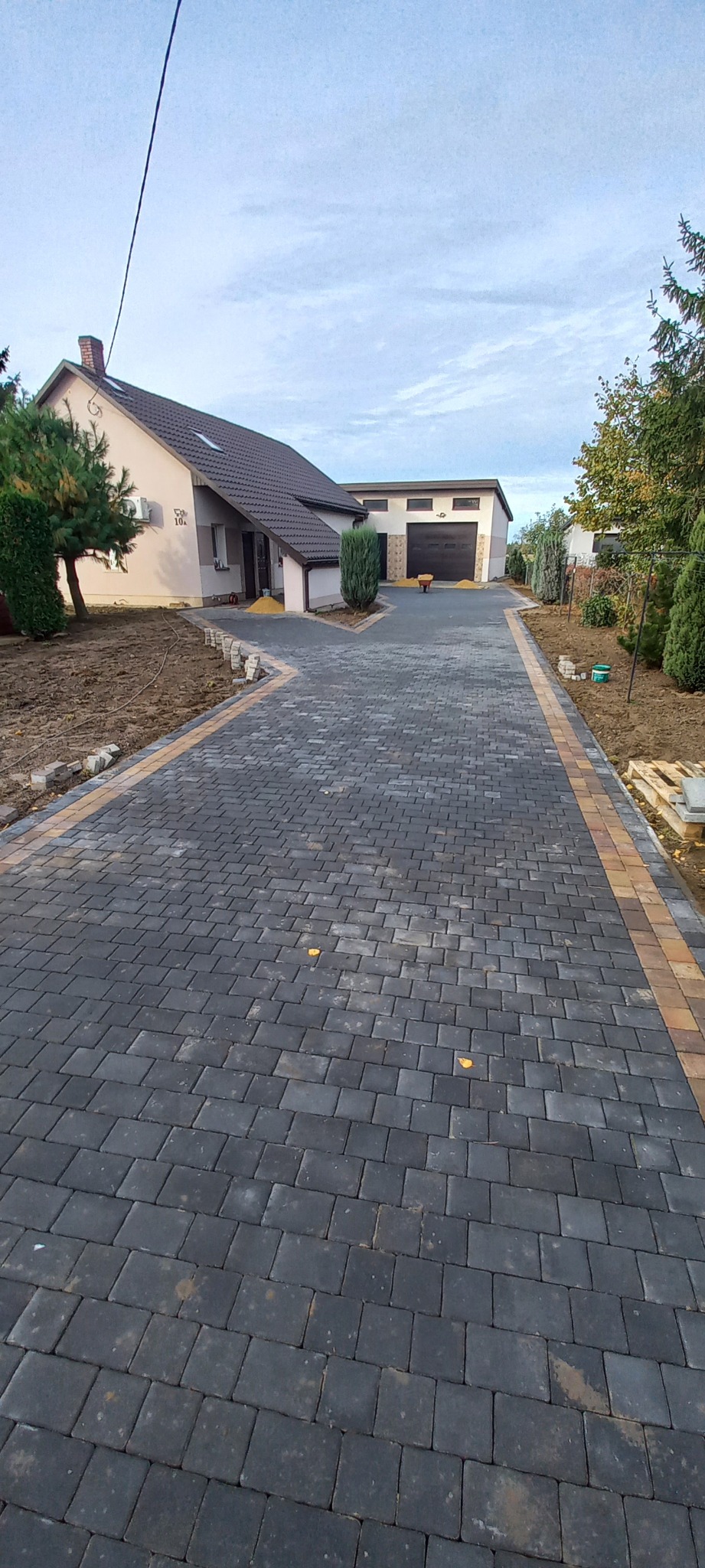 Ułożona, ciemnoszara kostka brukowa z obramowaniem w kolorze piaskowym prowadząca do domu i garażu. Częściowo przygotowane rabaty wokół.
