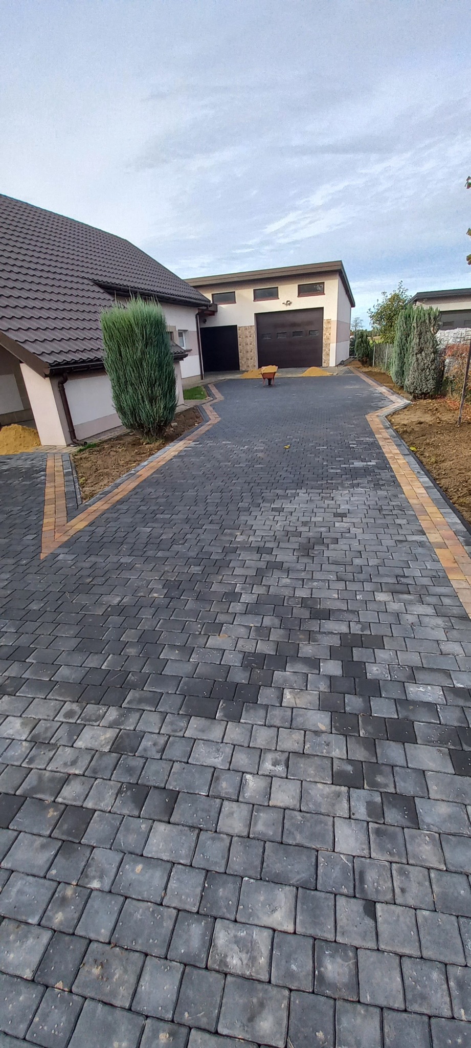Ułożona, ciemnoszara kostka brukowa z obramowaniem z jaśniejszych kostek, prowadząca do garażu z dwiema bramami; w tle wózek budowlany z piaskiem.
