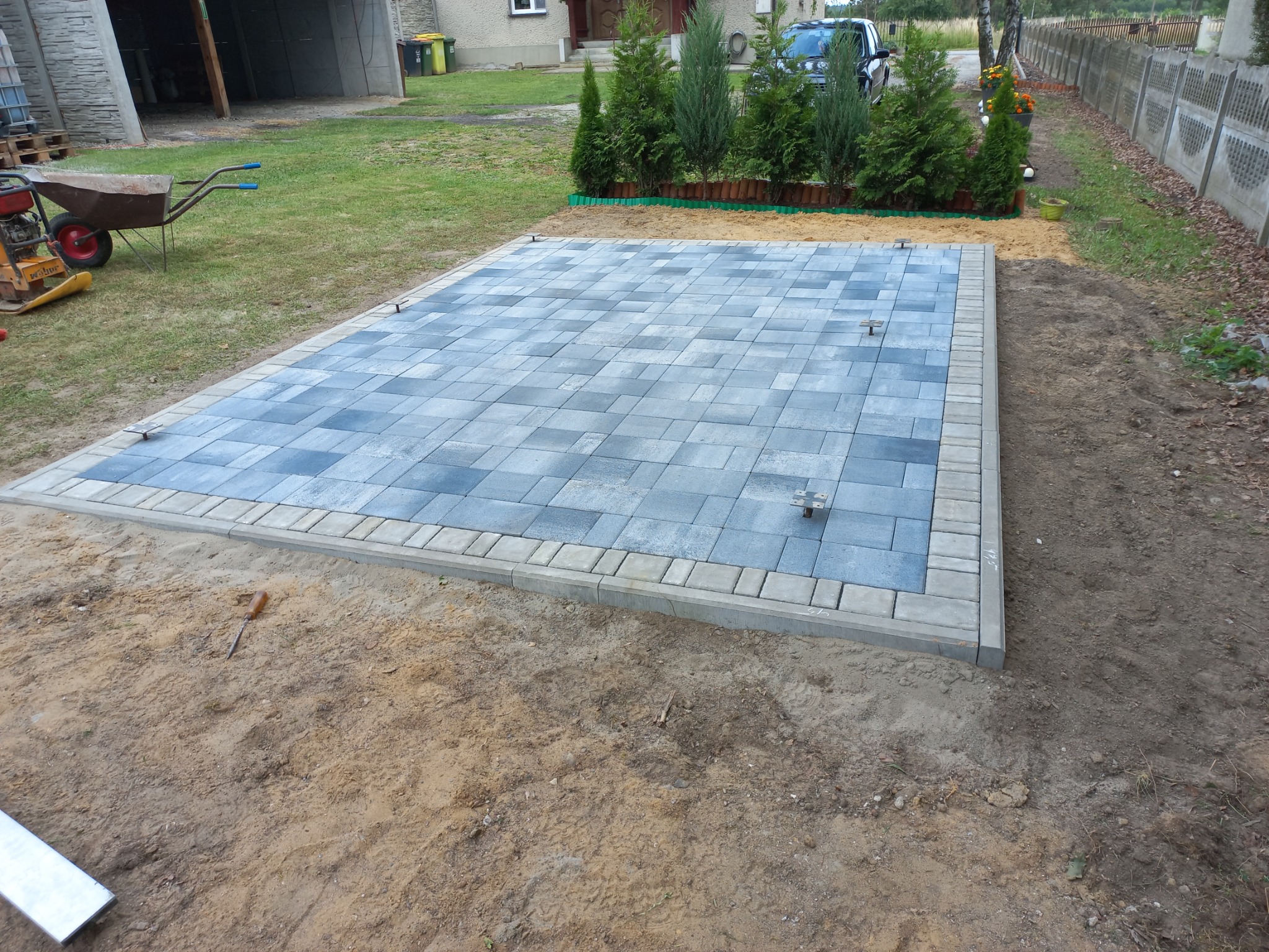 Powierzchnia z ułożonej szarej kostki brukowej z obrzeżem, przygotowana pod budowę, widoczne wystające metalowe elementy mocujące, w tle trawa, drzewa iglaste i betonowe ogrodzenie.