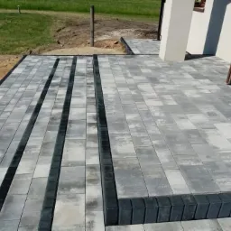 Ułożona szara kostka brukowa z ciemnymi pasami oddzielającymi, prowadząca do wejścia domu z białymi filarami i brązowymi drzwiami, widoczna trawa w tle.