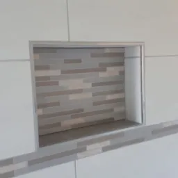 Wnęka w ścianie wyłożona mozaiką z płytek w odcieniach szarości i beżu, obramowana metalową listwą, otoczona białymi płytkami z szarymi akcentami wzdłuż krawędzi.