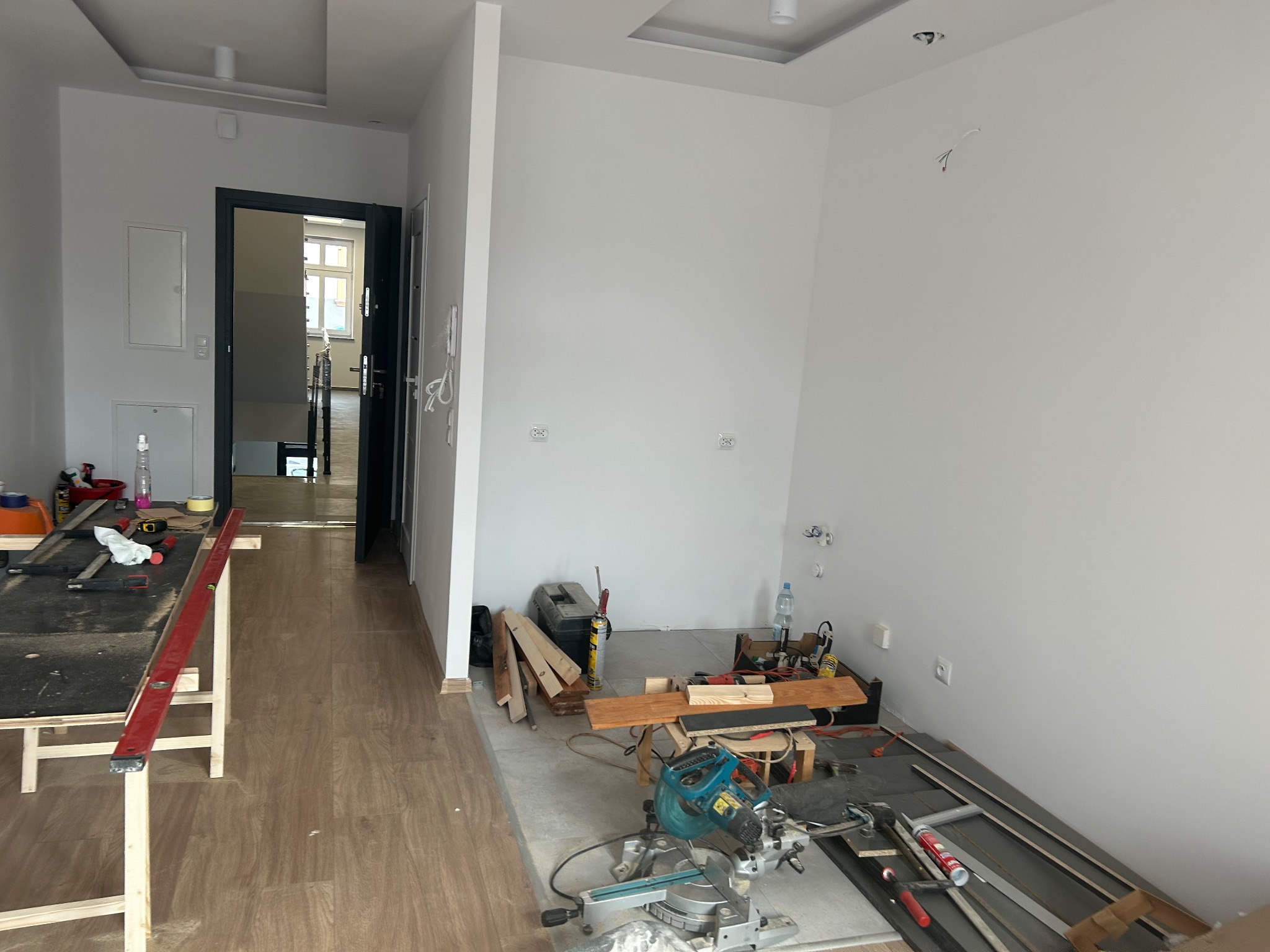 Przygotowania do prac wykończeniowych w pustym pomieszczeniu: widoczny stół roboczy z narzędziami, poziomica, piła ukośnica, skrzynka narzędziowa i materiały budowlane na podłodze.