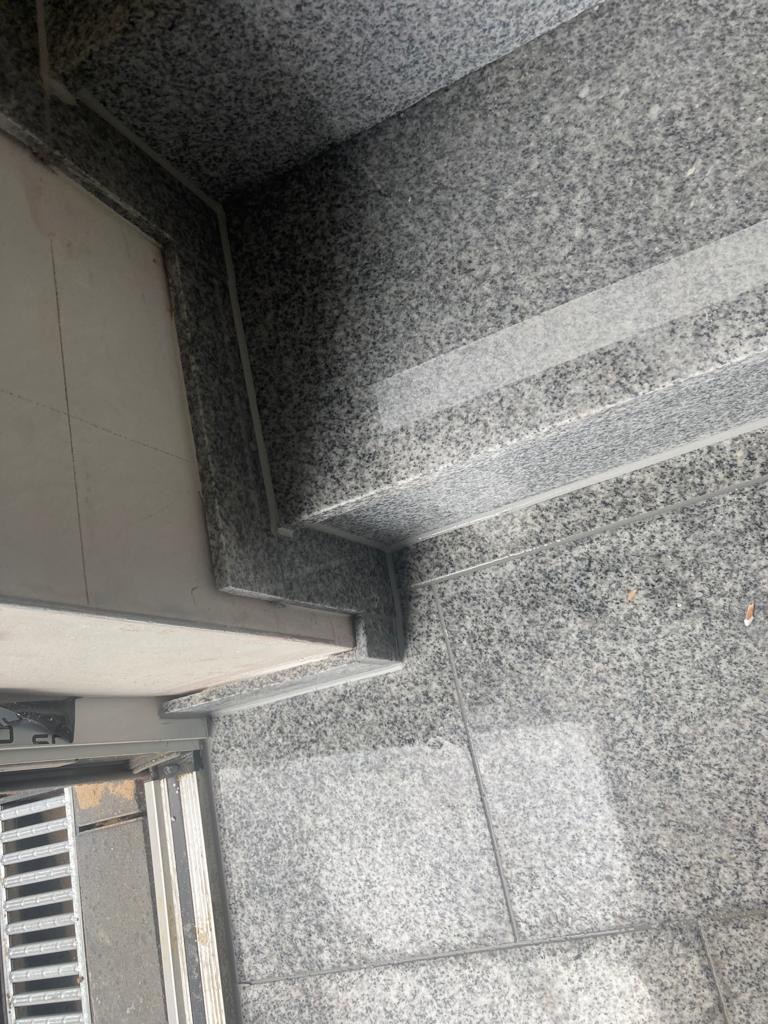 Granitowe schody wejściowe z szarymi płytkami podłogowymi, widoczne z bliska, z metalową kratką w tle