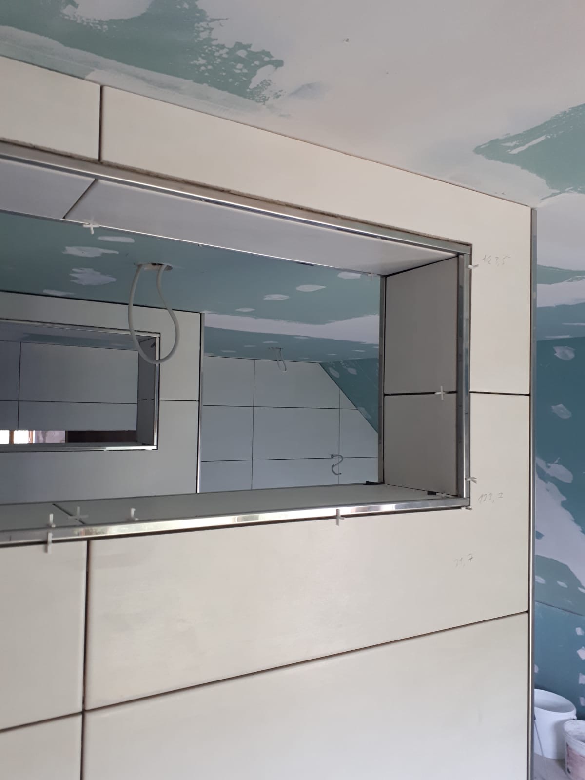 Świeżo ułożone, jasne płytki ceramiczne w łazience z wnęką wykończoną metalową listwą. Widoczne krzyżyki dystansowe i notatki na ścianie. W odbiciu widać dalszą część łazienki z białymi płytkami.