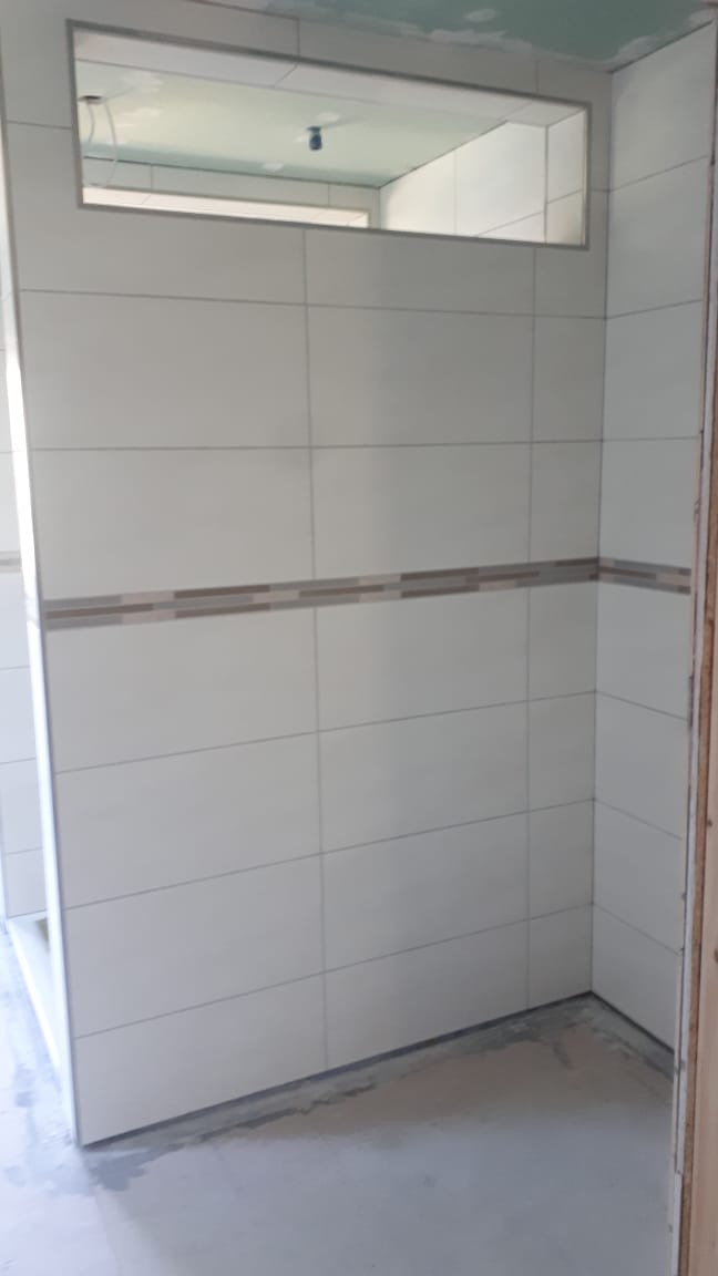 Wnętrze łazienki w trakcie remontu: białe płytki ceramiczne z dekoracyjnym pasem, surowy beton na podłodze, widoczny otwór w ścianie z lustrzanym odbiciem dalszej części pomieszczenia, sufit...