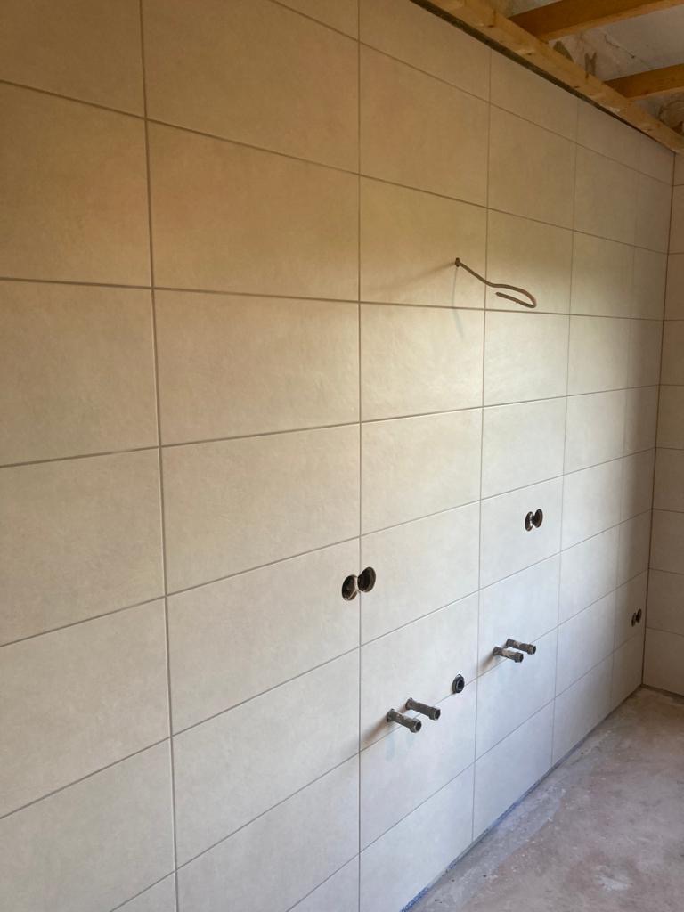 Ściana wyłożona jasnymi, kwadratowymi płytkami ceramicznymi z widocznymi otworami na instalacje wodne i elektryczne, w trakcie remontu łazienki.
