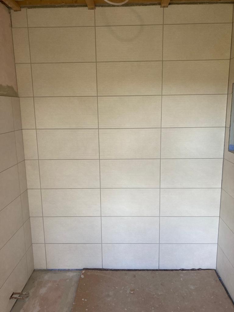 Ściana łazienki wyłożona beżowymi płytkami z poziomymi paskami, widoczne niedokończone fugowanie i surowa podłoga.