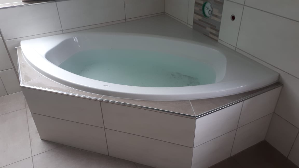 Narożna wanna wypełniona wodą, obudowana jasnymi, kwadratowymi płytkami ceramicznymi z szarą fugą, wykończona srebrną listwą, na ścianie białe płytki z szarym akcentem dekoracyjnym i zegarem.