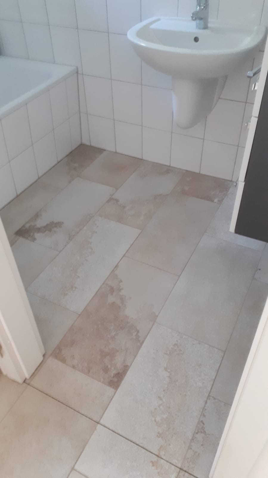 Jasne, prostokątne płytki podłogowe w łazience z umywalką wiszącą i fragmentem wanny, widoczne delikatne, nieregularne wzory na płytkach.