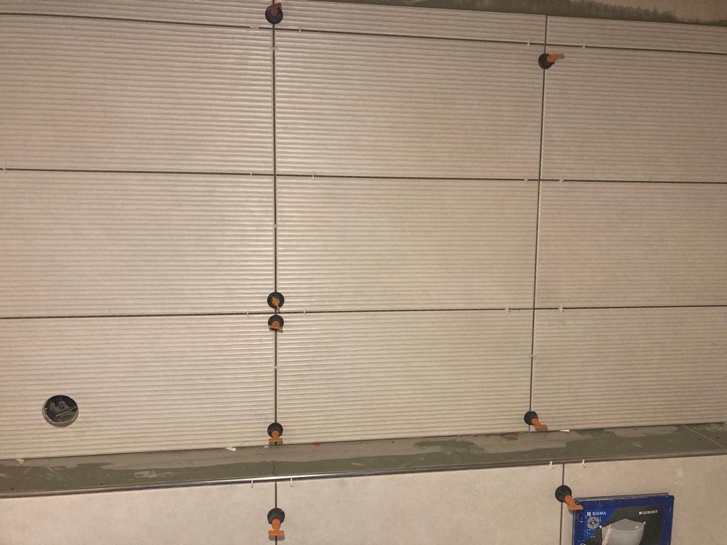 Montaż płytek ściennych z użyciem poziomowania krzyżykowego; widoczne płytki z delikatnym prążkowanym wzorem i otwór na instalację.