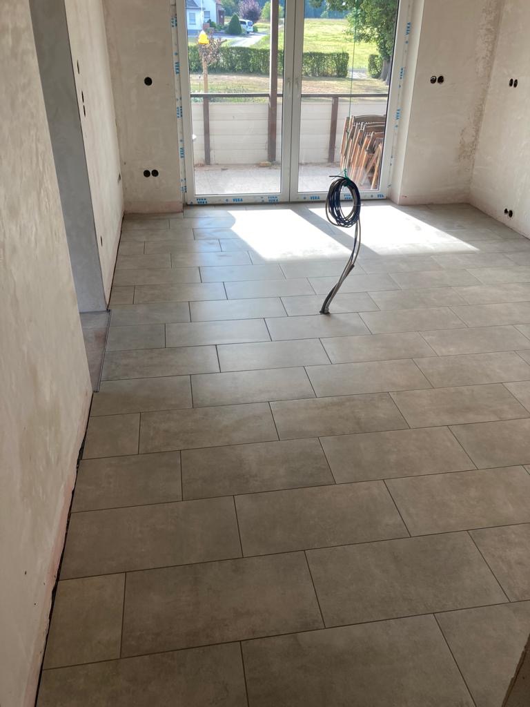 Ułożone szare, prostokątne płytki podłogowe w jasnym pomieszczeniu z widocznymi niedokończonymi ścianami i kablem wystającym z podłogi w pobliżu okna balkonowego.