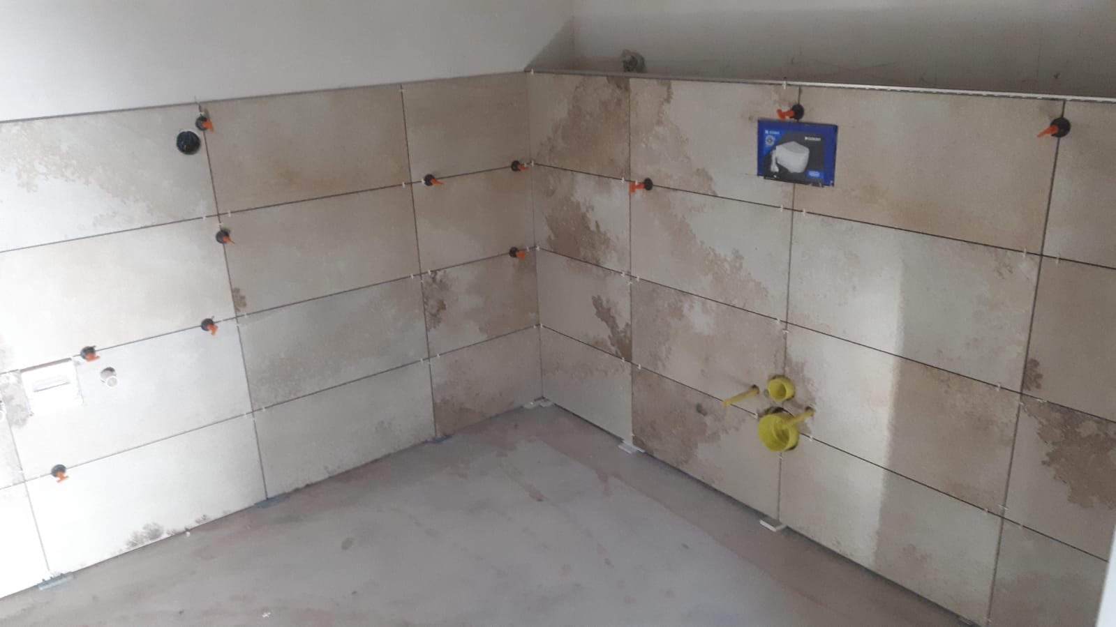 Ściana łazienki w trakcie układania beżowych płytek z wzorem, z widocznymi krzyżykami dystansowymi i elementami instalacji wodnej w żółtej osłonie.